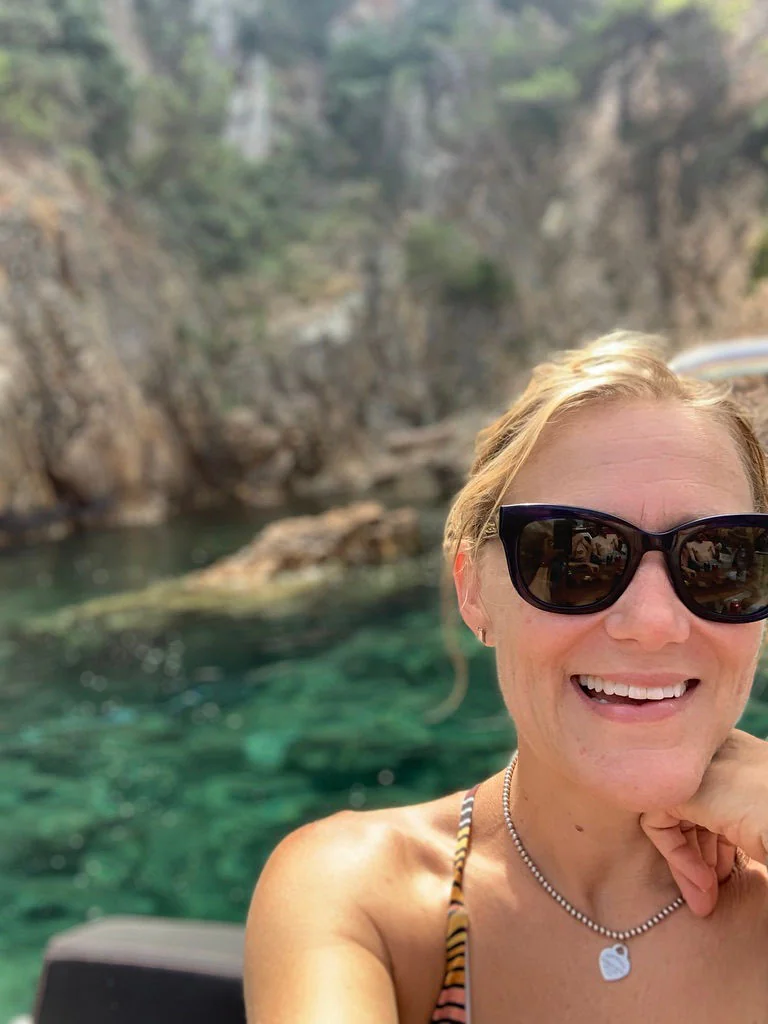 Woman smiling on a boat with turquoise Mediterranean water and rocky cliffs in the background.