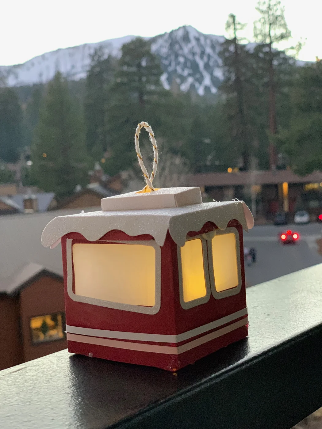 A lit paper gondola on a balcony rail at dusk, the village lights and mountain behind it