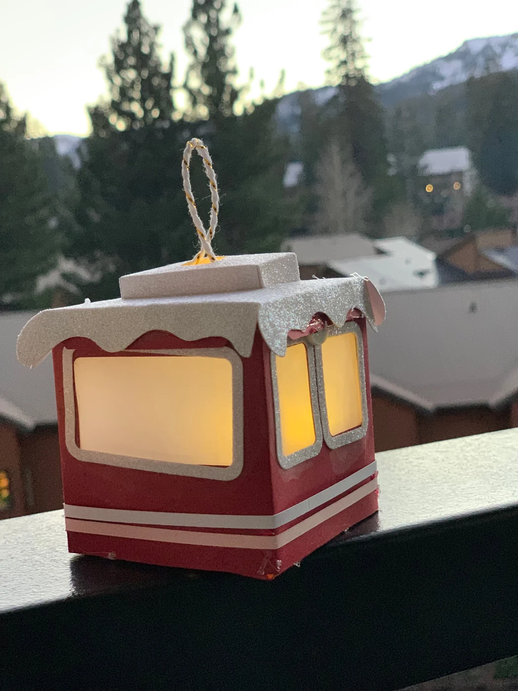 A paper gondola lantern with a snow-capped roof glowing on a balcony rail, snowy mountain behind it