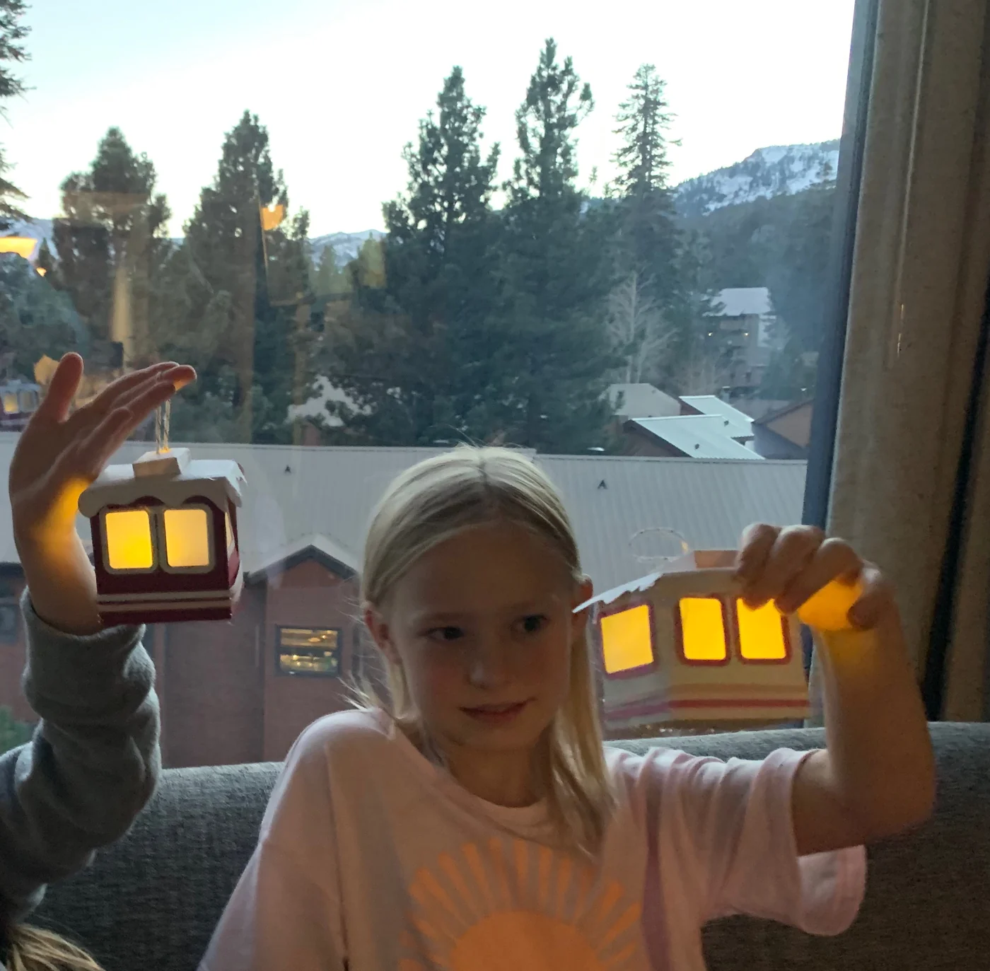 A girl holding up two glowing paper gondola lanterns in a hotel room at dusk