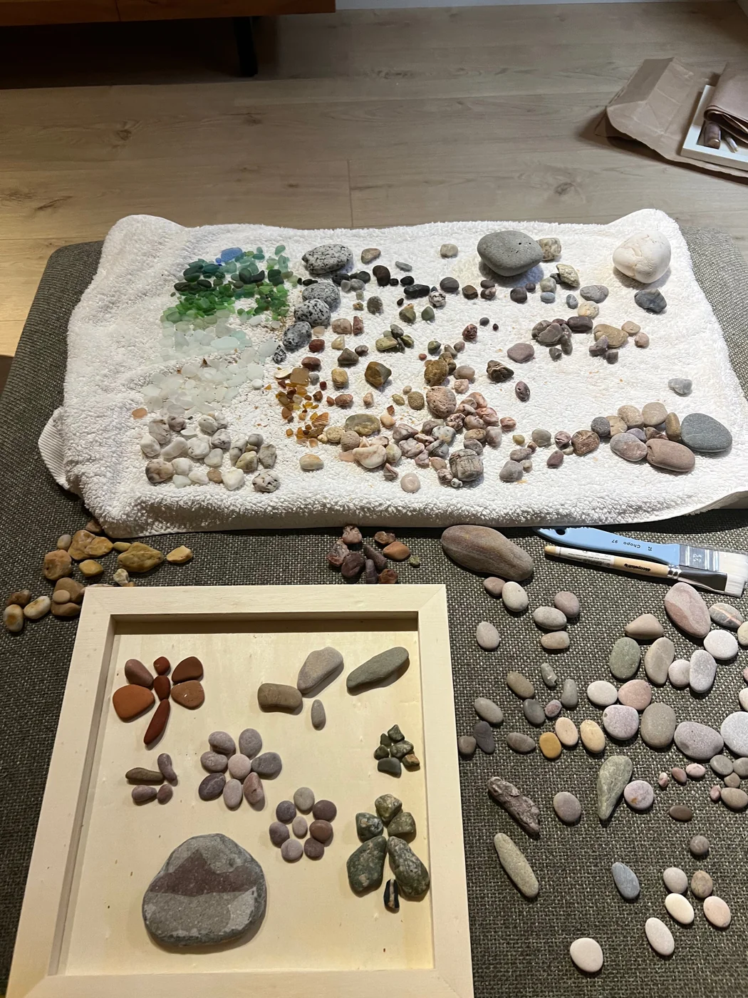 The home sorting station: pebbles arranged by color on a towel beside a white frame holding a half-made pebble mosaic