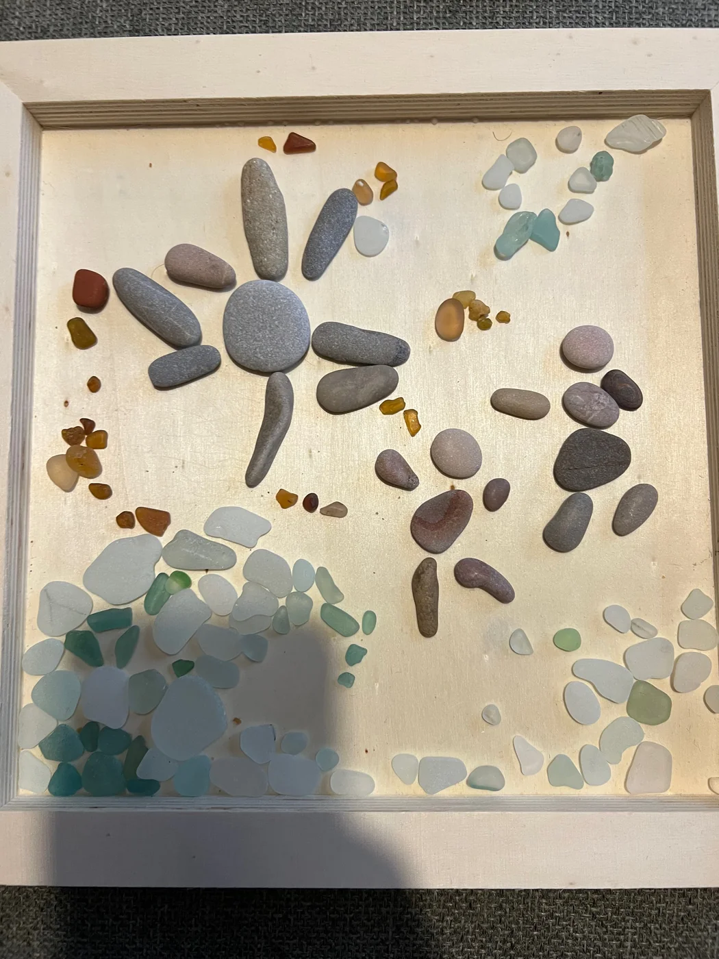 A white frame holding a mosaic of pebbles and sea glass arranged into a sun and scattered blooms