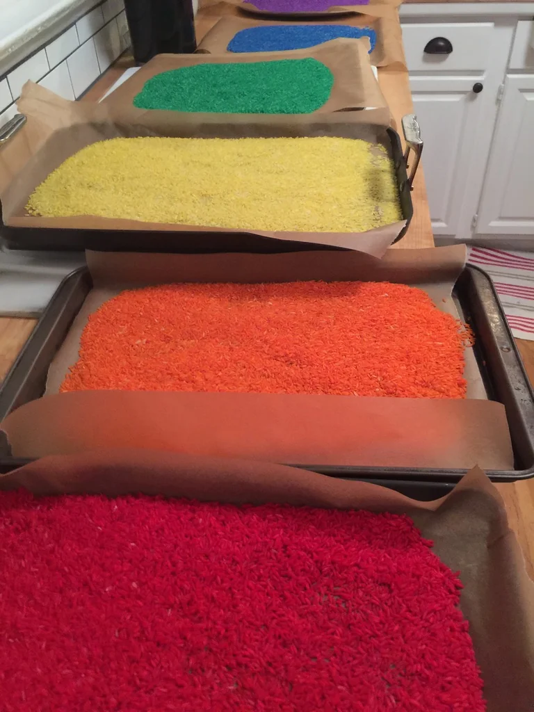 Trays of red, orange, yellow, green, blue, and purple rice drying on parchment-lined baking sheets