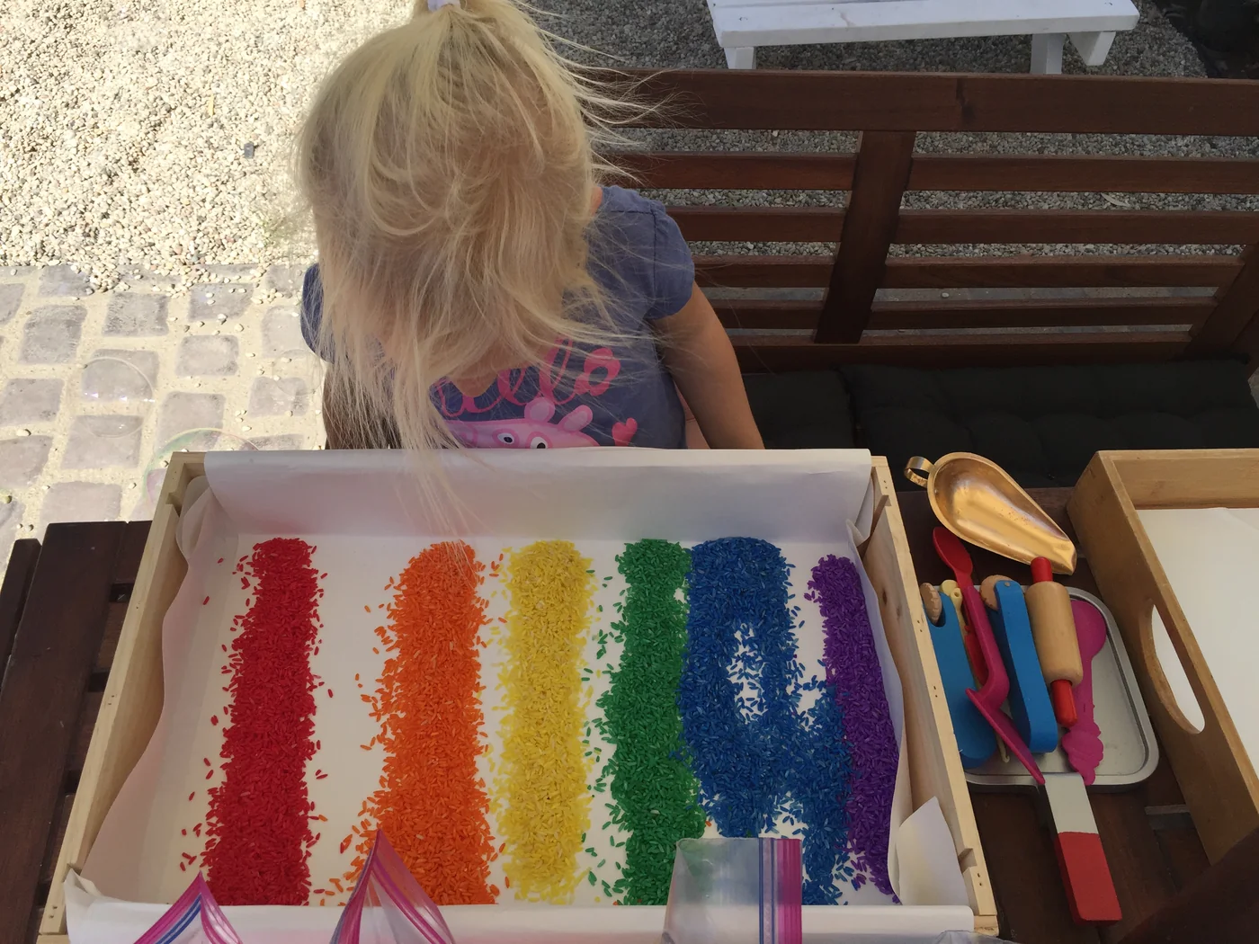 A child playing at a wooden tray filled with six bright stripes of rainbow-colored rice
