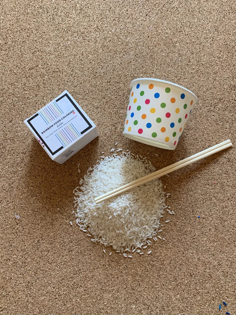 Everything you need for rainbow rice: a box of rainbow food coloring, a polka dot paper cup, a pile of white rice, and takeout chopsticks