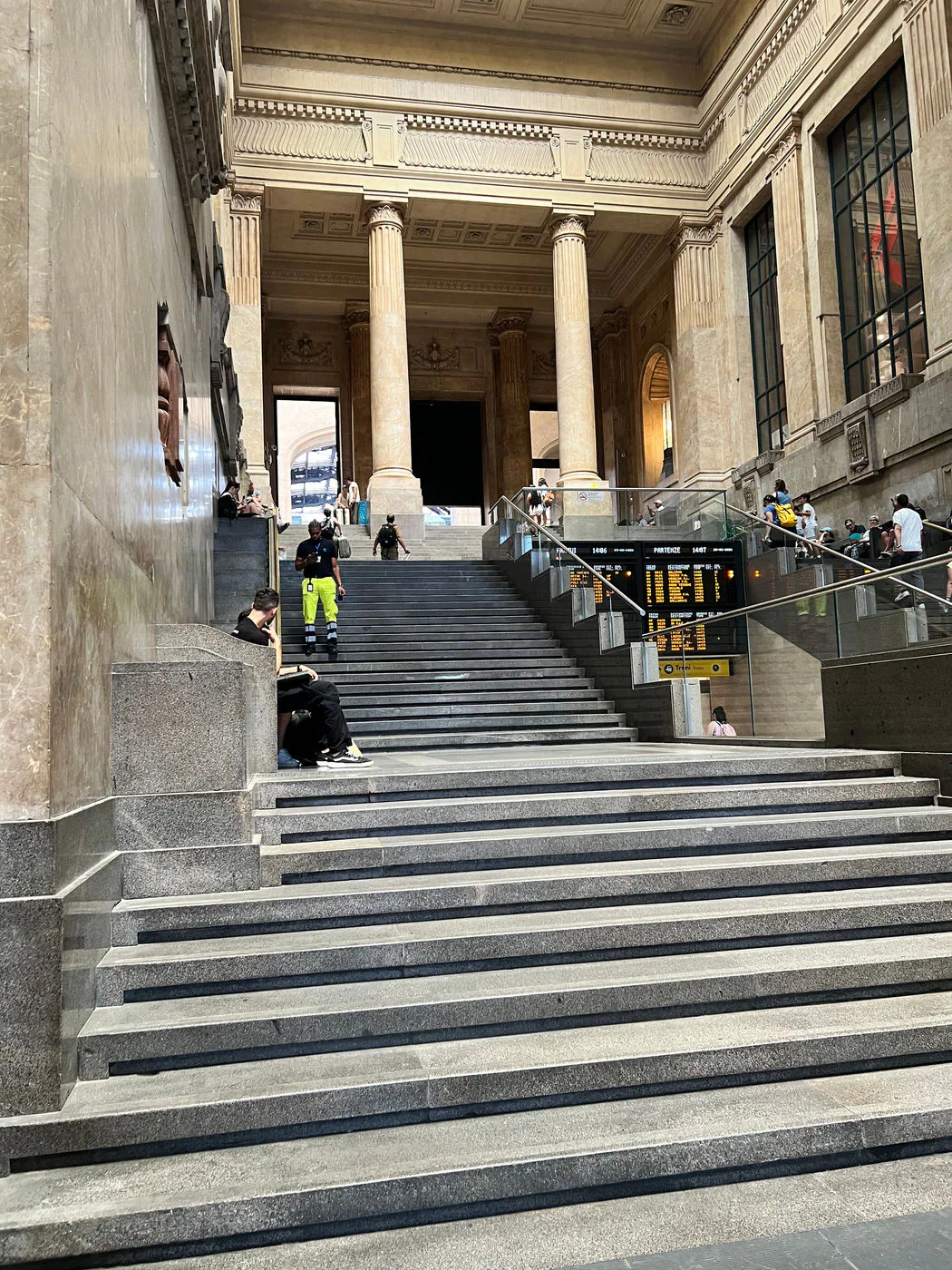 The wide marble staircase inside Milan Centrale station beneath tall columns and departure boards.