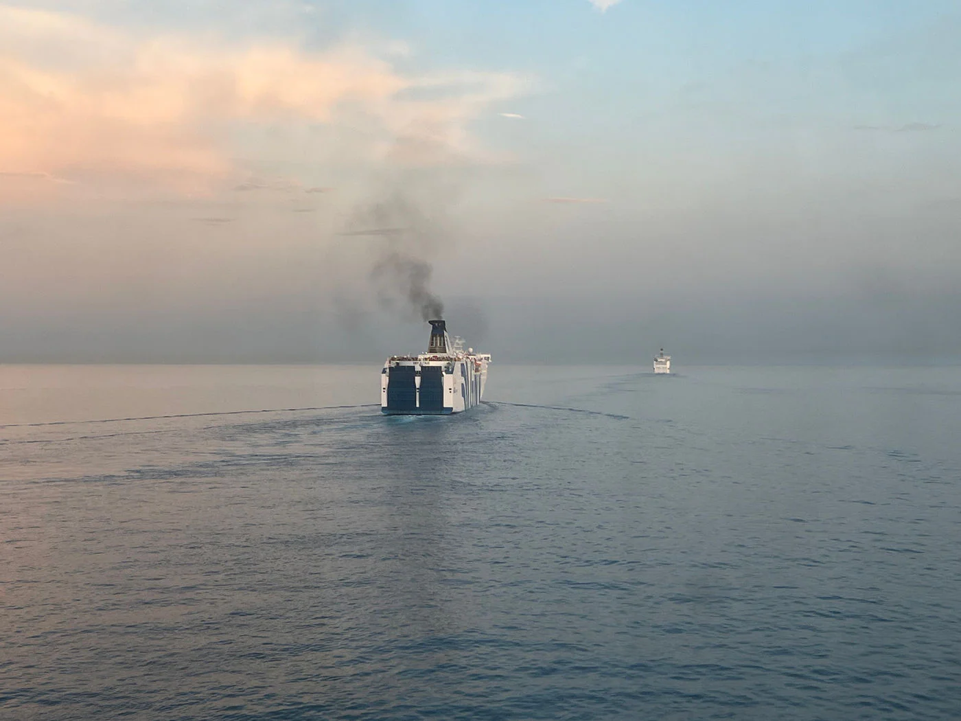A large ferry sailing ahead across a calm sea at sunset, a second ship on the horizon.