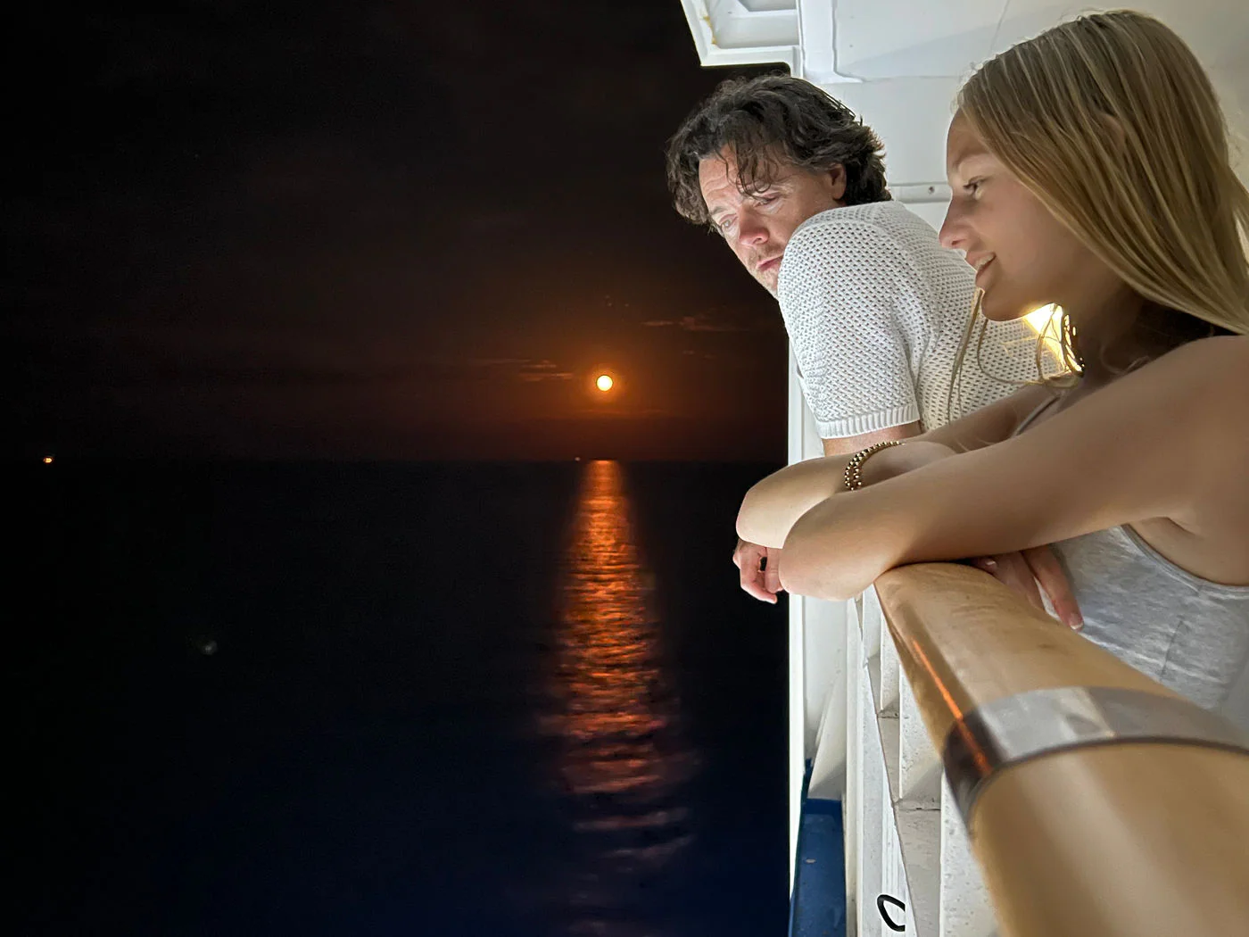 Two passengers lean on a ship's railing at night watching the moon's reflection on dark water.