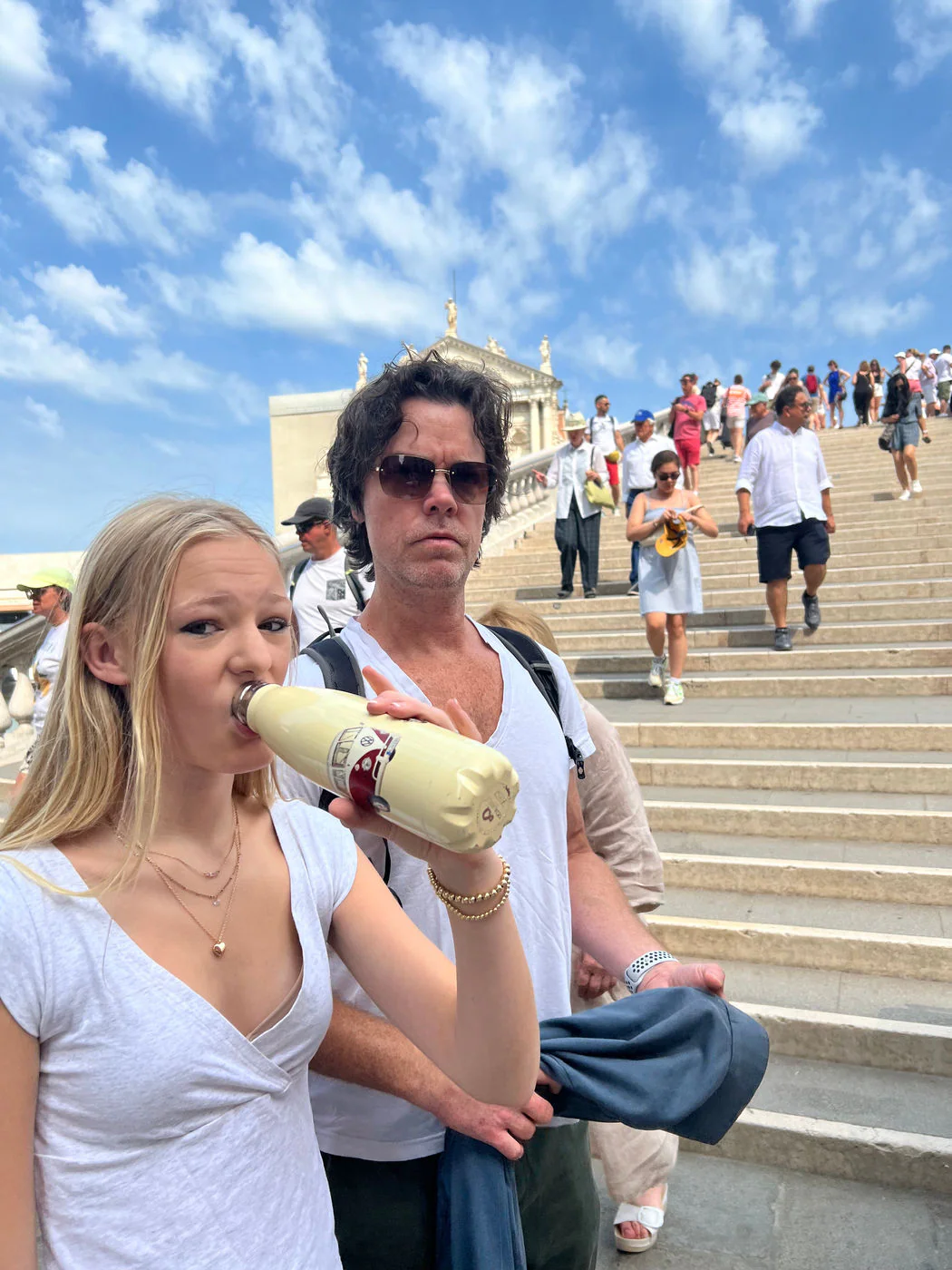 A traveler drinks from a water bottle on sunlit stone steps while another shelters from the heat behind.