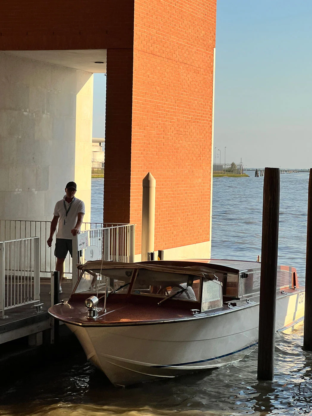A wooden water taxi moored at a covered airport dock beside a tall brick pillar, a staff member standing at the gate.